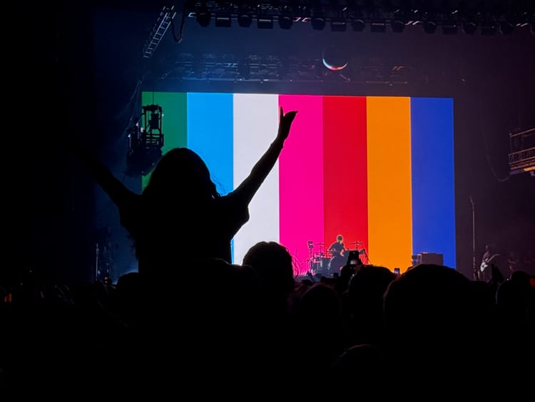 A silhouette of a person sitting on the shoulders of another person, watching the band Turnstile play in front of a technicolor screen. 